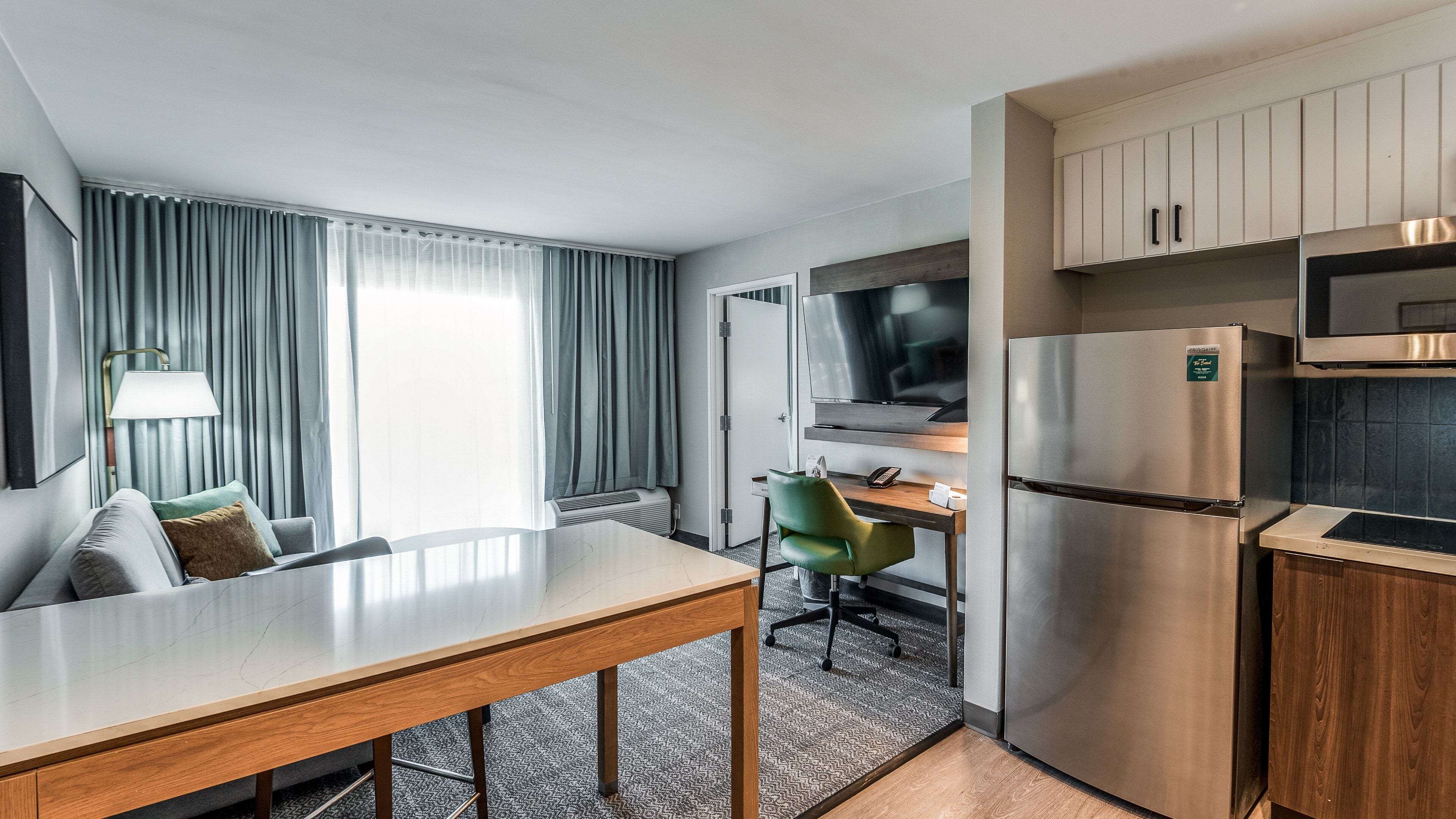 Une chambre d’hôtel compacte comprenant une table à manger, un canapé, un fauteuil vert, une télévision, un réfrigérateur et un four à micro-ondes.