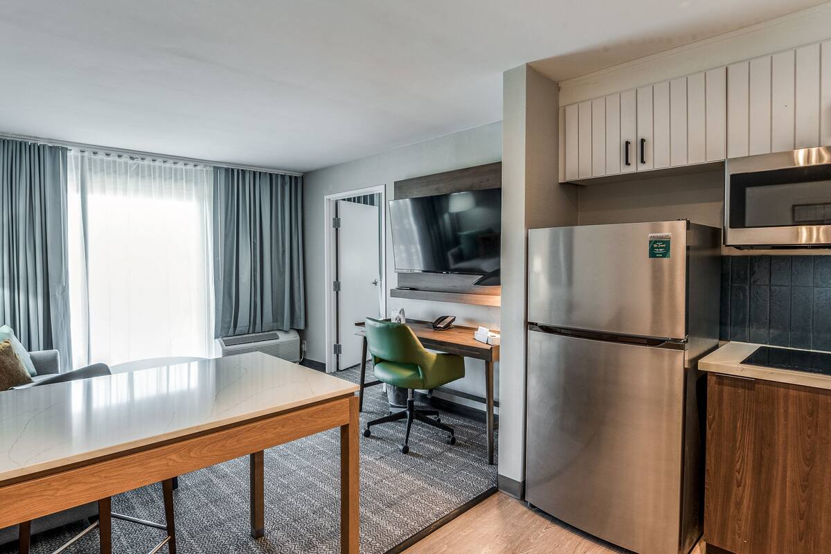 Una habitación de hotel compacta con una mesa de comedor, un sofá, una silla verde, un televisor, un refrigerador y un microondas.