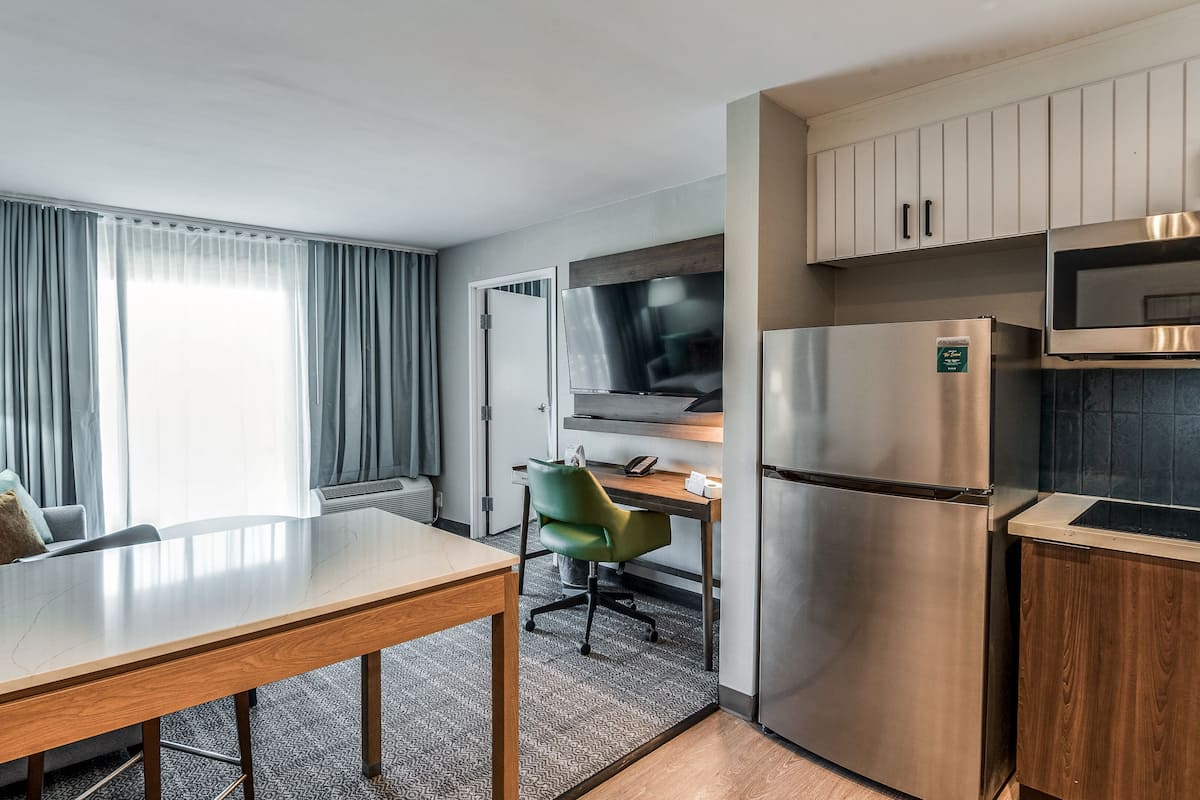 Une chambre d’hôtel compacte comprenant une table à manger, un canapé, un fauteuil vert, une télévision, un réfrigérateur et un four à micro-ondes.