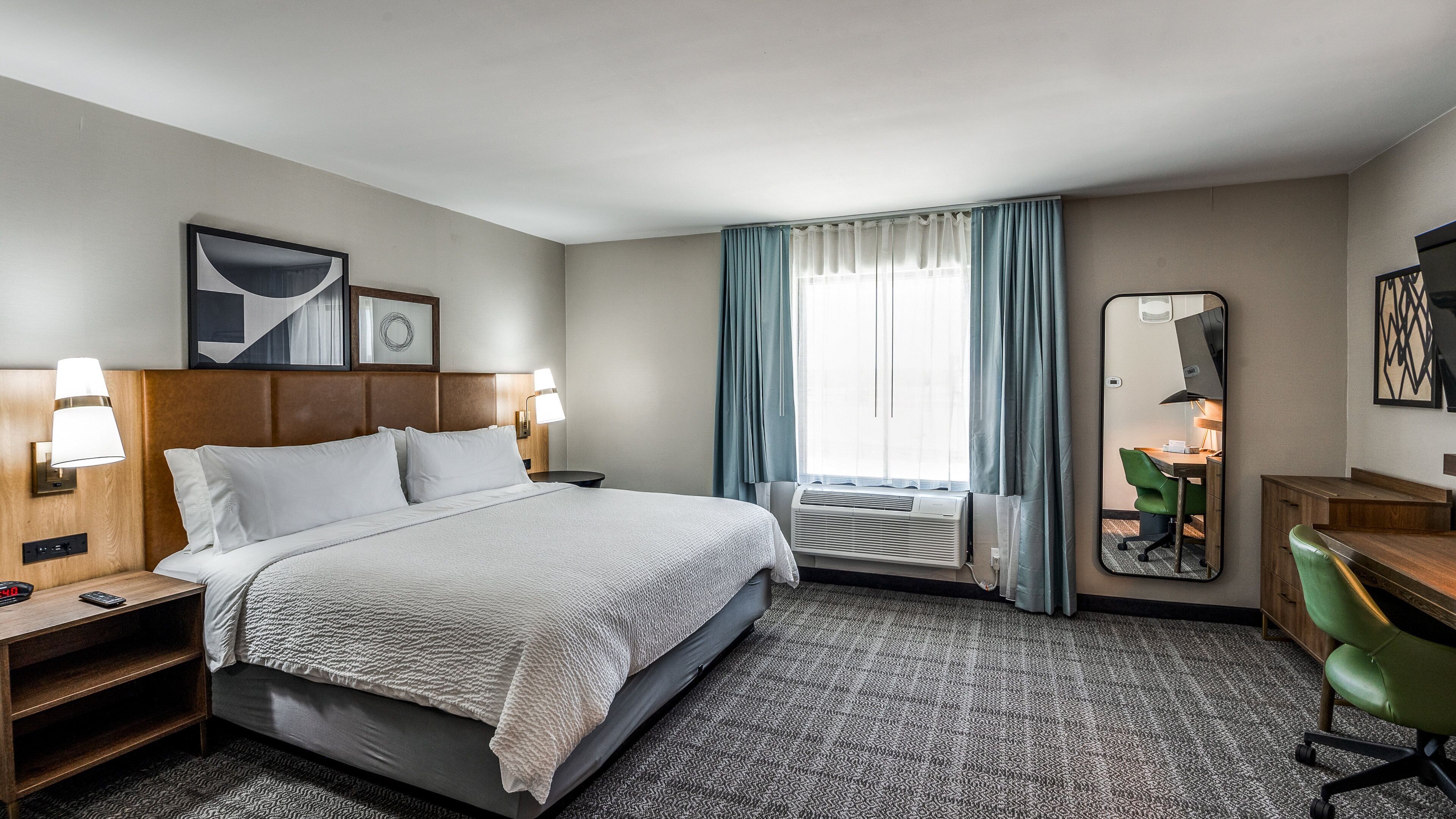 A hotel room with a large bed, bedside lamps, a desk with a chair, a mirror, and a window with curtains.