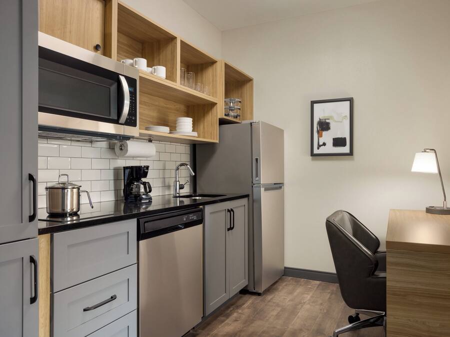 Eine kompakte Küche mit Mikrowelle, Kaffeemaschine und Kühlschrank. Darüber befindet sich ein Holzregal und darunter ein kleiner Tisch mit einer Lampe.