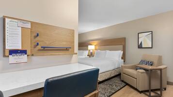 A hotel room with a bed, a desk, a chair, a small table, a wall-mounted bulletin board, and a framed picture.