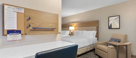 A hotel room with a bed, a desk, a chair, a small table, a wall-mounted bulletin board, and a framed picture.