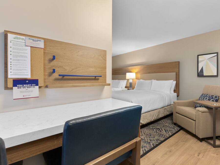 A hotel room with a bed, a desk, a chair, a small table, a wall-mounted bulletin board, and a framed picture.