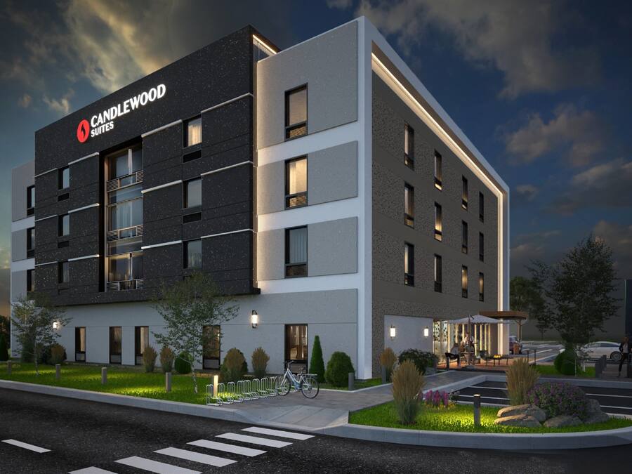 Un hotel moderno con un letrero que dice "CANDLEWOOD SUITES", que presenta una combinación de fachada oscura y clara, varios balcones y un área de entrada ajardinada.