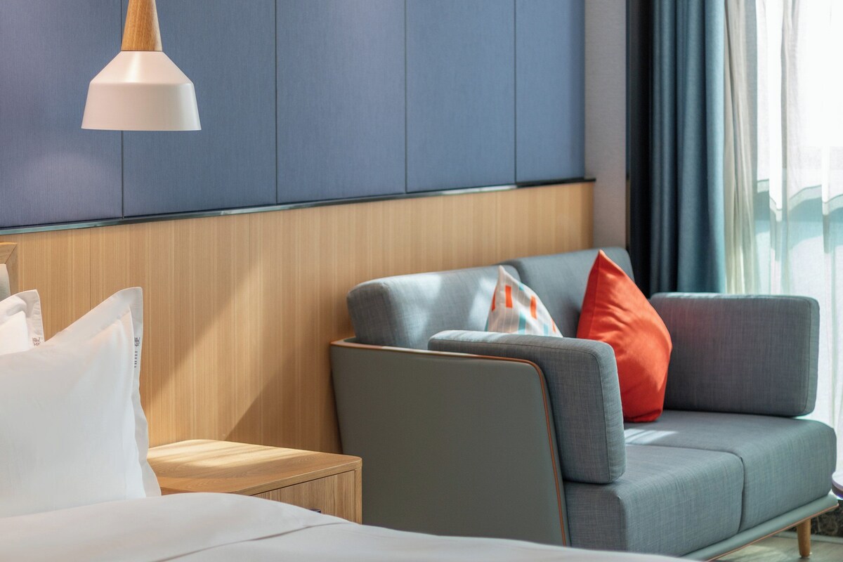 A hotel room with a bed, a grey sofa with red and white pillows, a wooden nightstand, and a white pendant light.