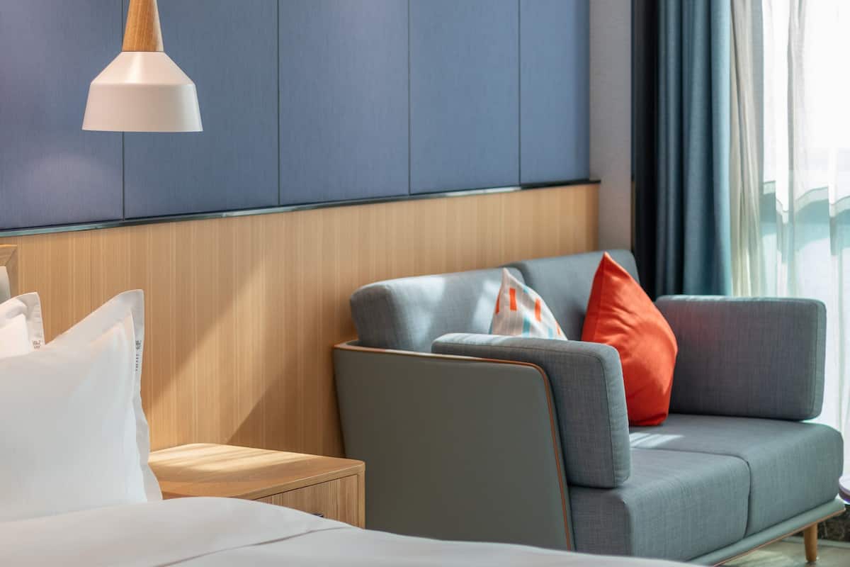 A hotel room with a bed, a grey sofa with red and white pillows, a wooden nightstand, and a white pendant light.
