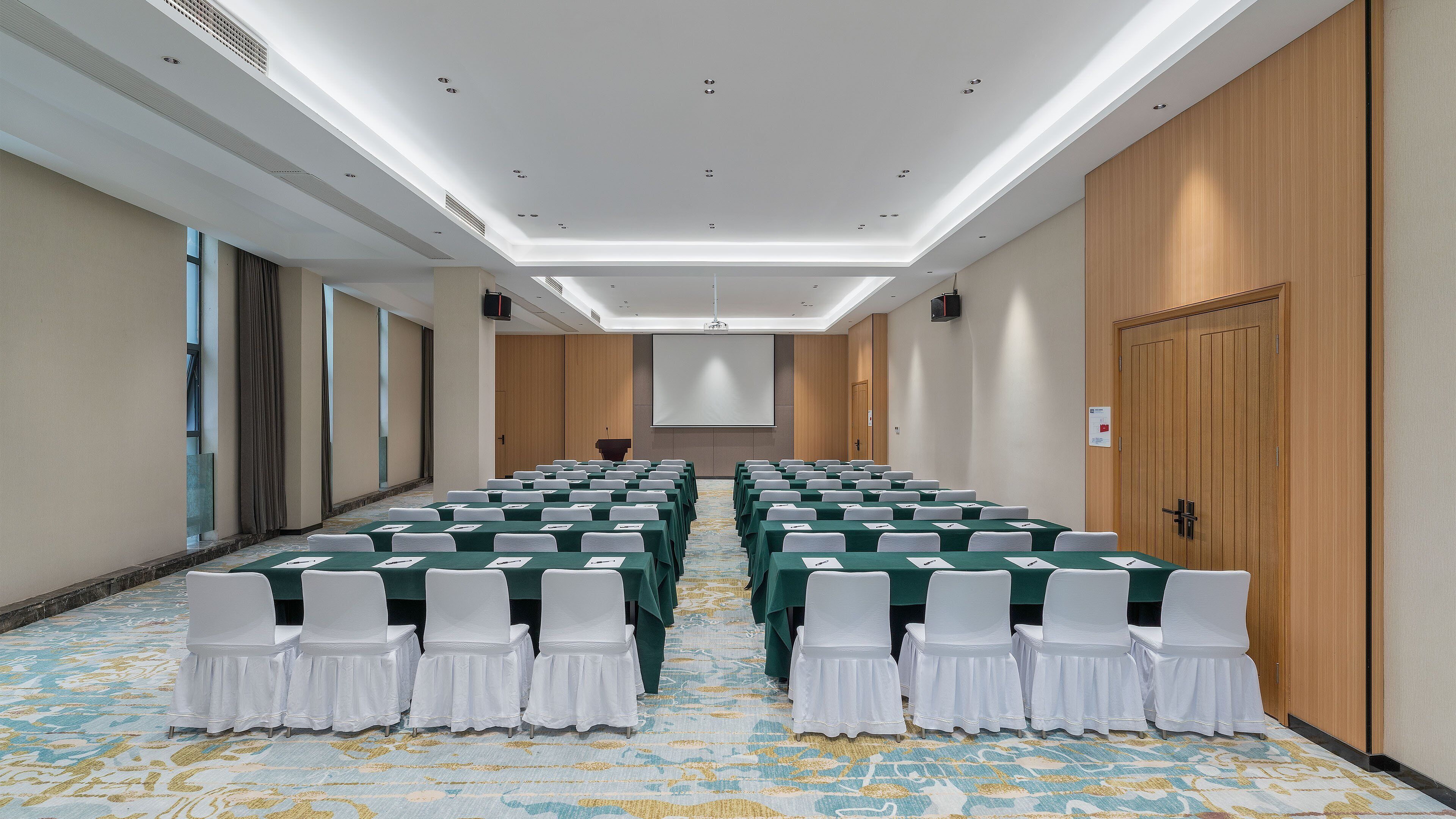 A conference room with rows of tables and chairs, a projector screen, and a carpeted floor.