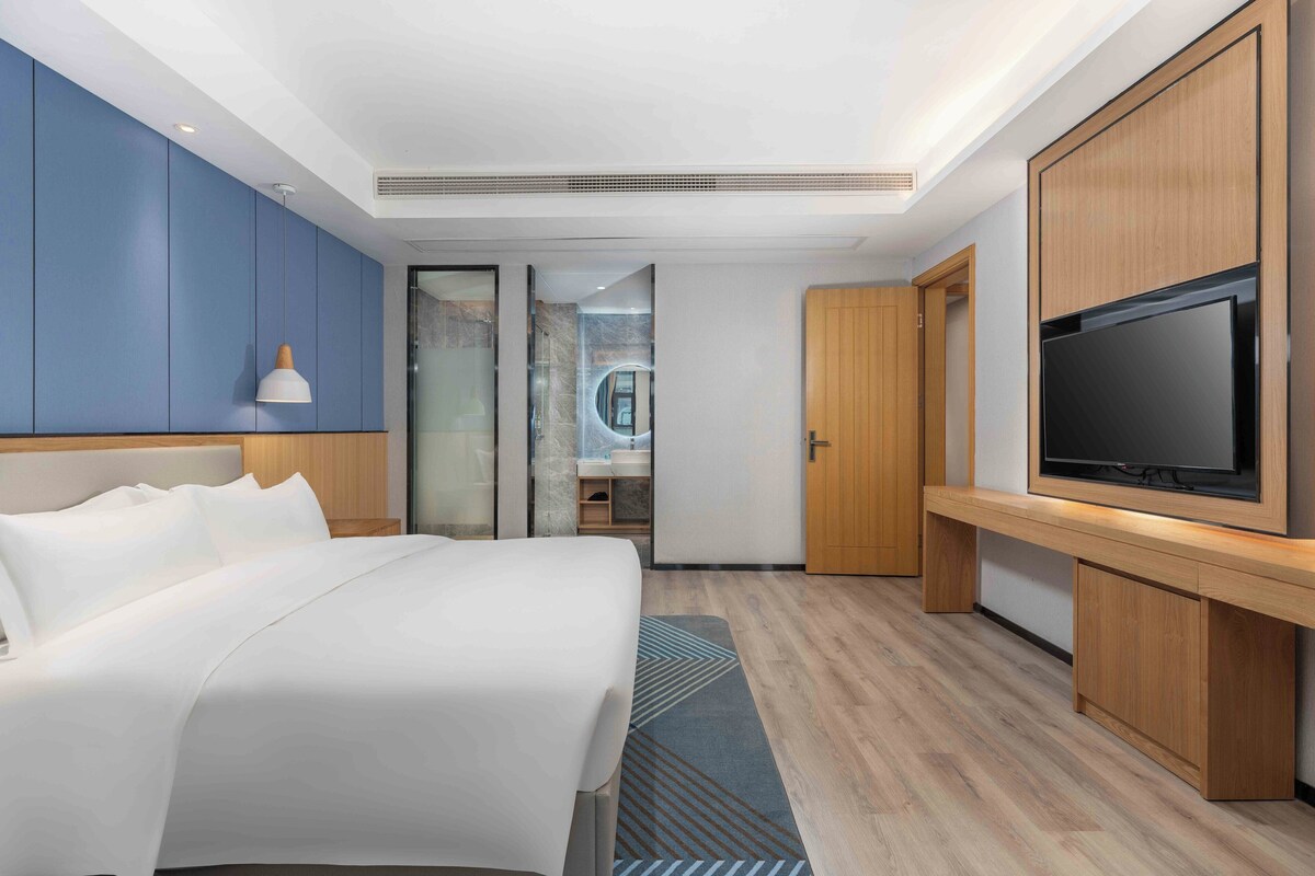 A modern hotel room with a large bed, a TV, wooden cabinets, and a bathroom visible through a glass door.