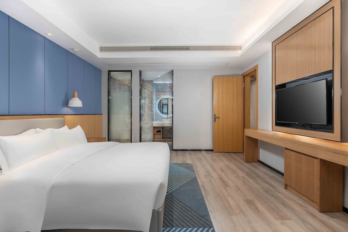 A modern hotel room with a large bed, a TV, wooden cabinets, and a bathroom visible through a glass door.
