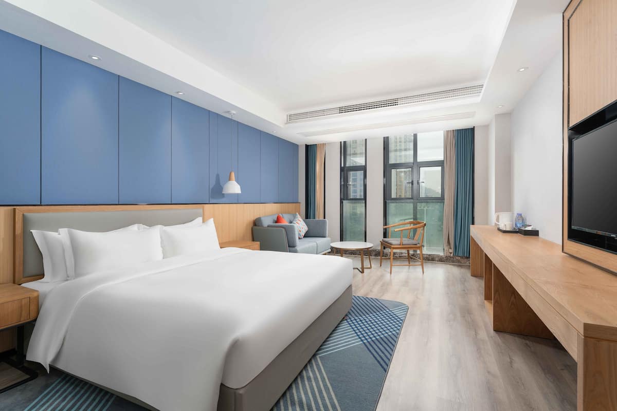 A modern hotel room with a large bed, a desk, a TV, and a view of the city through large windows.