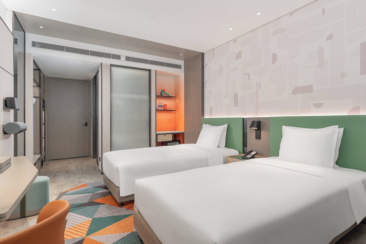 A modern hotel room with two beds, a desk, a chair, and a small table with a lamp.