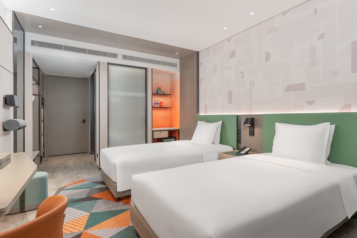 Ein modernes Hotelzimmer mit zwei Betten, einem Schreibtisch, einem Sessel und einem kleinen Tisch mit einer Lampe.