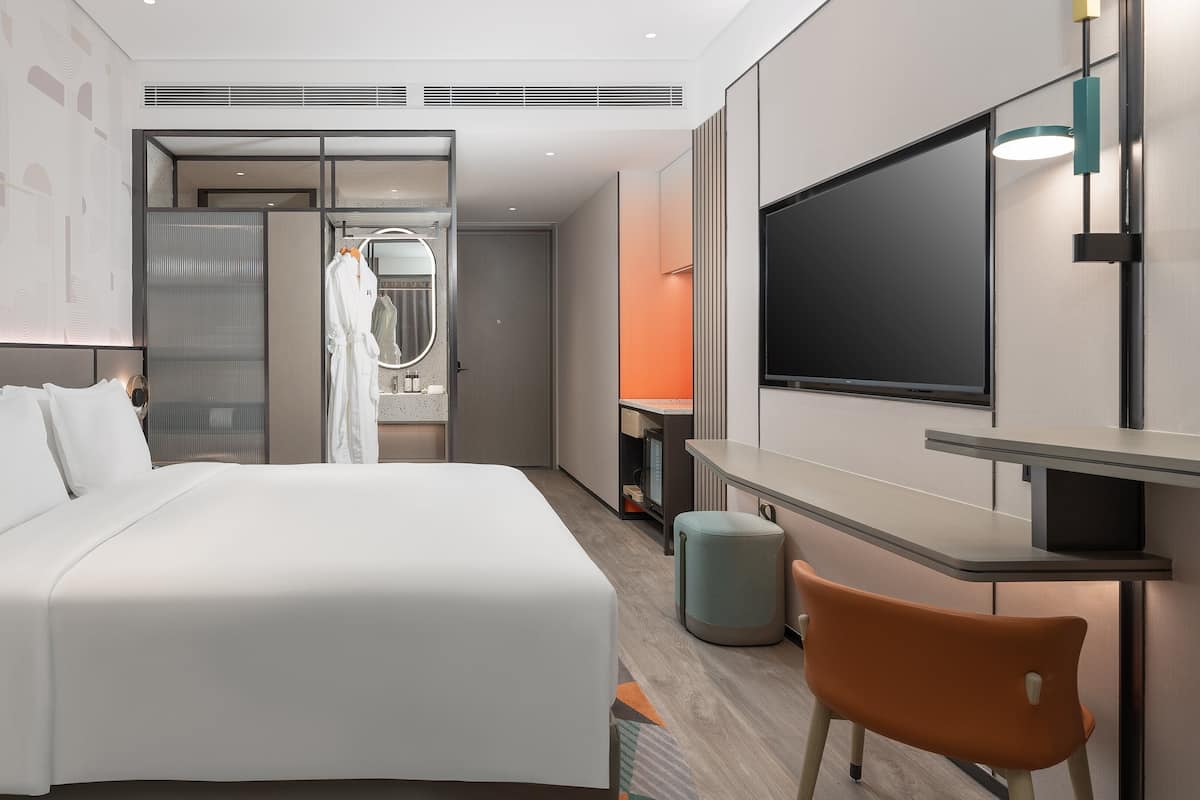 Ein modernes Hotelzimmer mit einem großen Bett, einem Fernseher, einem Schreibtisch und einem durch eine Glastrennwand sichtbaren Badezimmer.