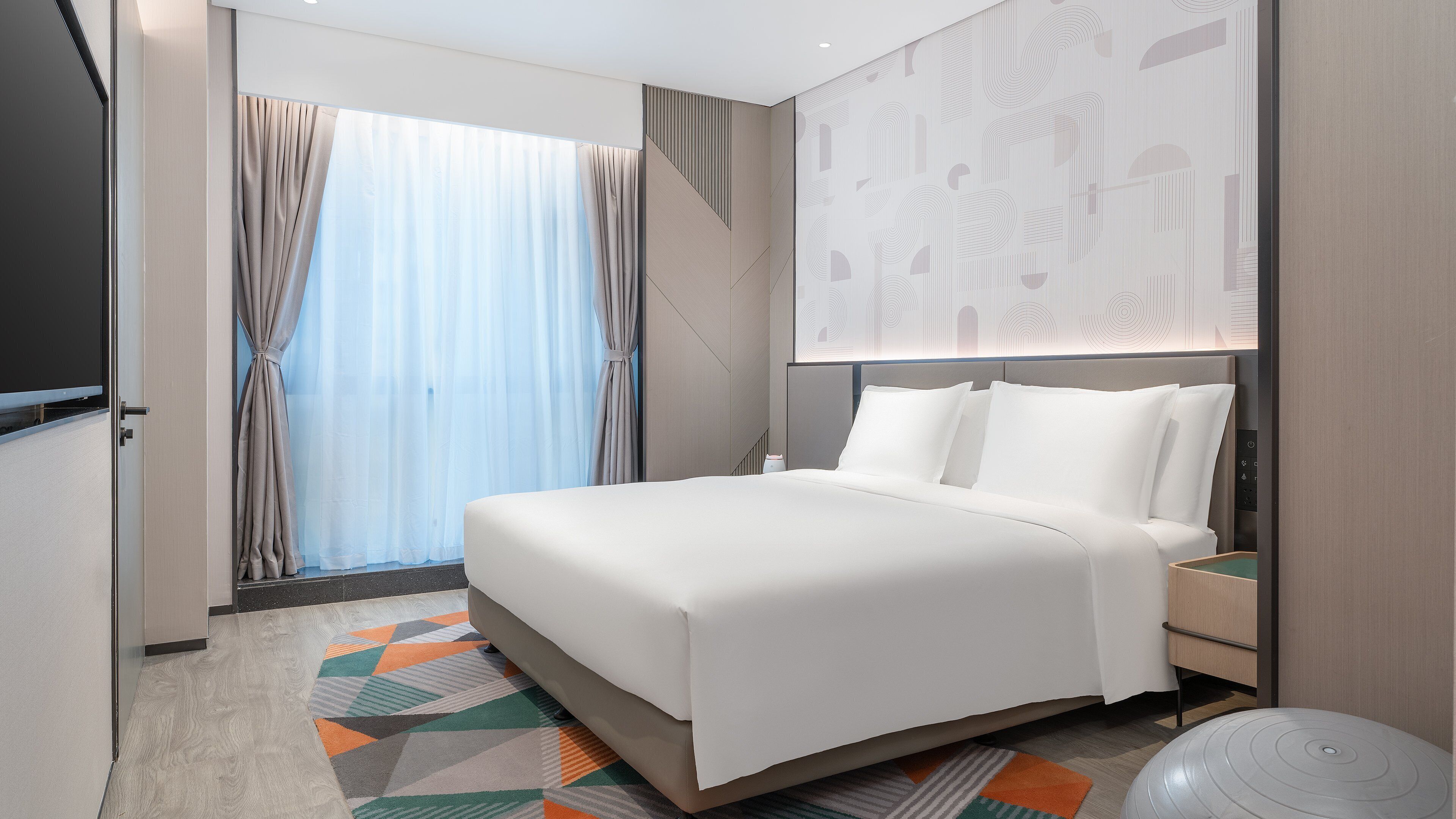 Una habitación de hotel moderna con una cama grande, una mesita de noche, un televisor montado en la pared y un ventanal con cortinas.