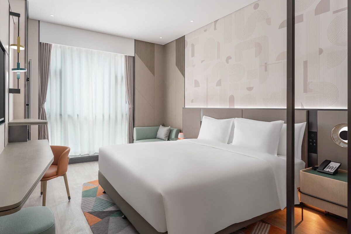 A modern hotel room with a bed, desk, chair, and a small table with a phone.