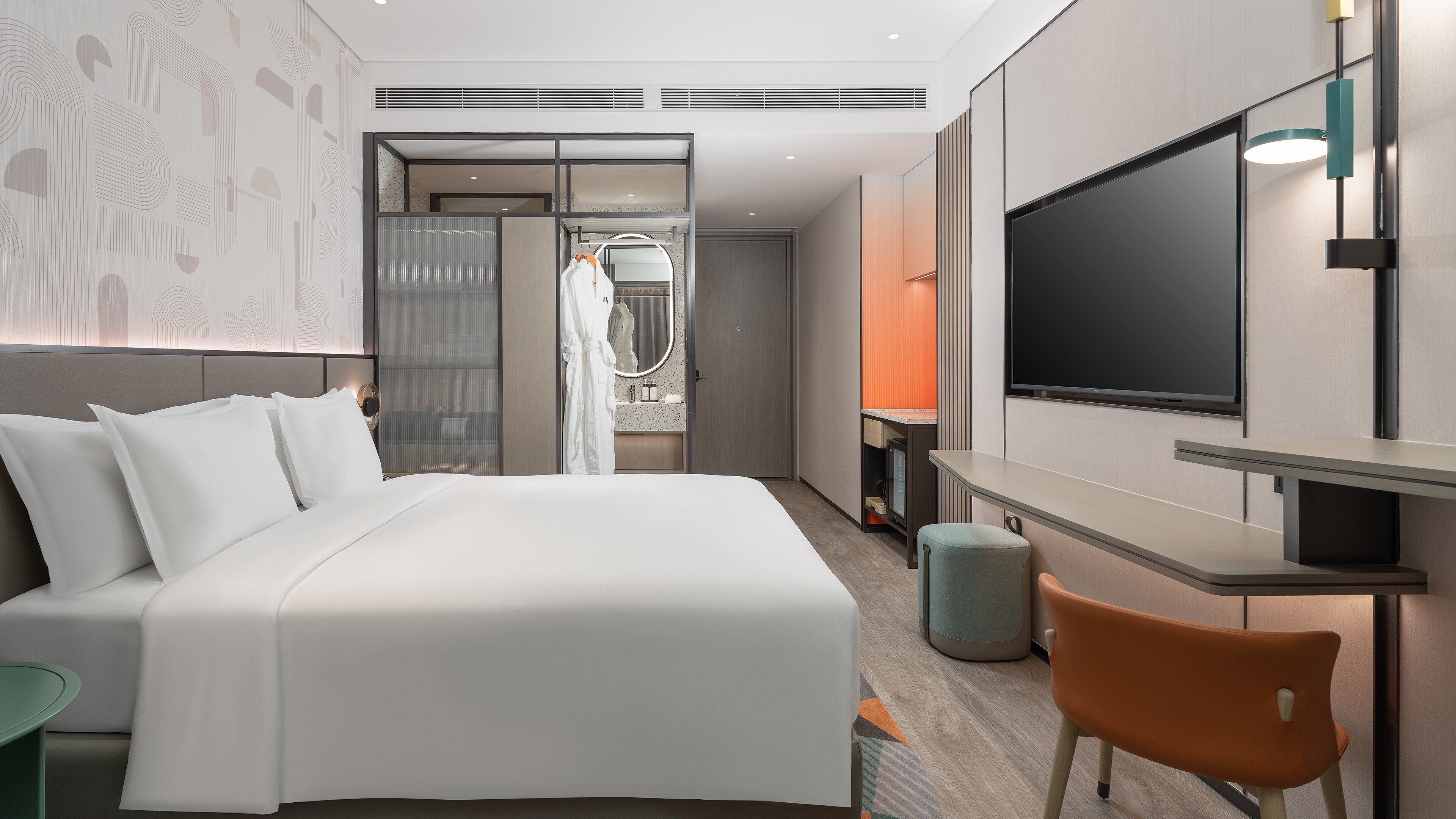 A modern hotel room with a large bed, a TV, a desk, and a bathroom visible through a glass partition.