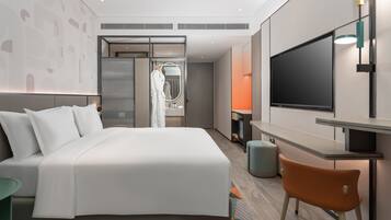 A modern hotel room with a large bed, a TV, a desk, and a bathroom visible through a glass partition.