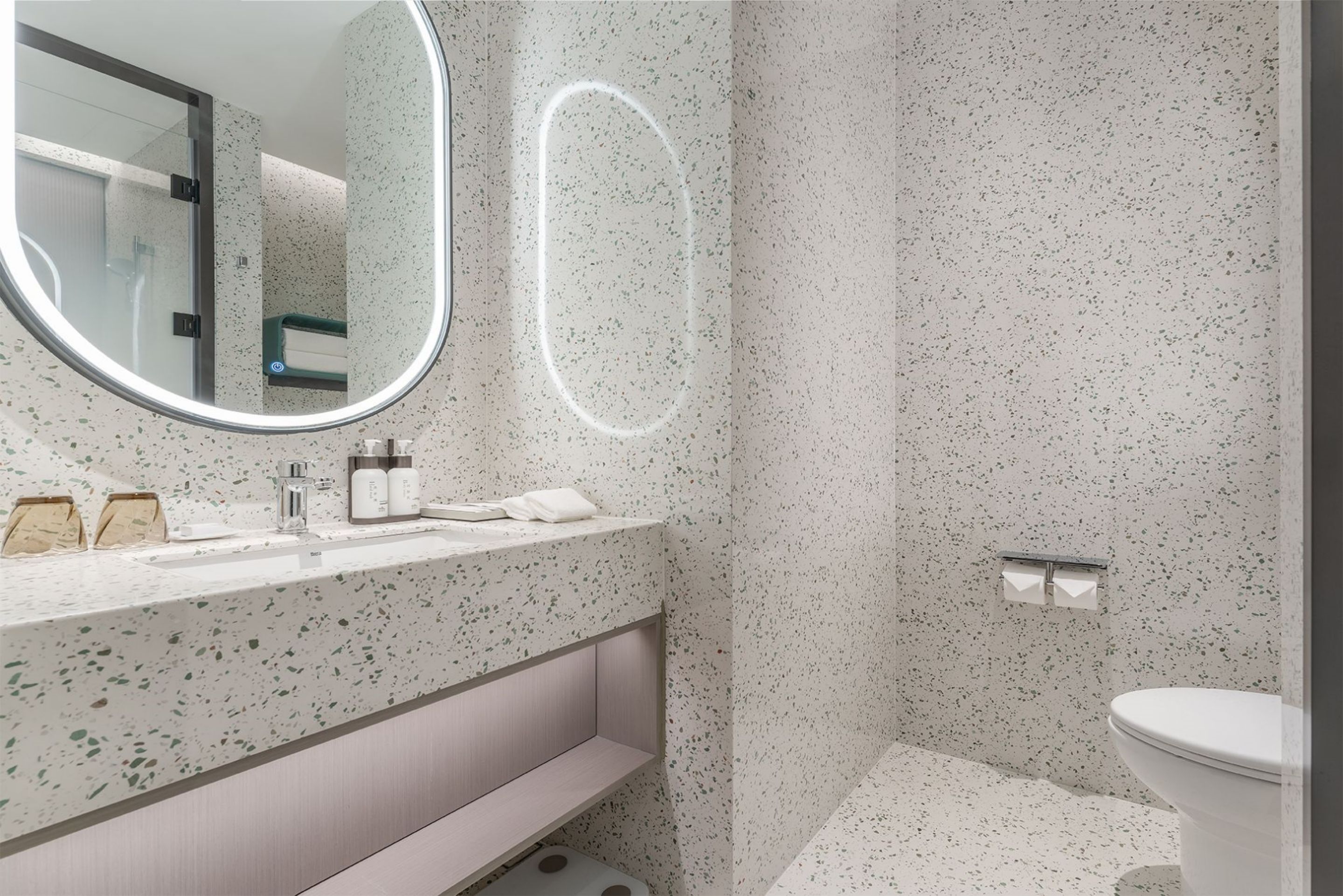 A modern bathroom with a speckled backsplash, a round mirror with a light, a sink with toiletries, and a toilet.