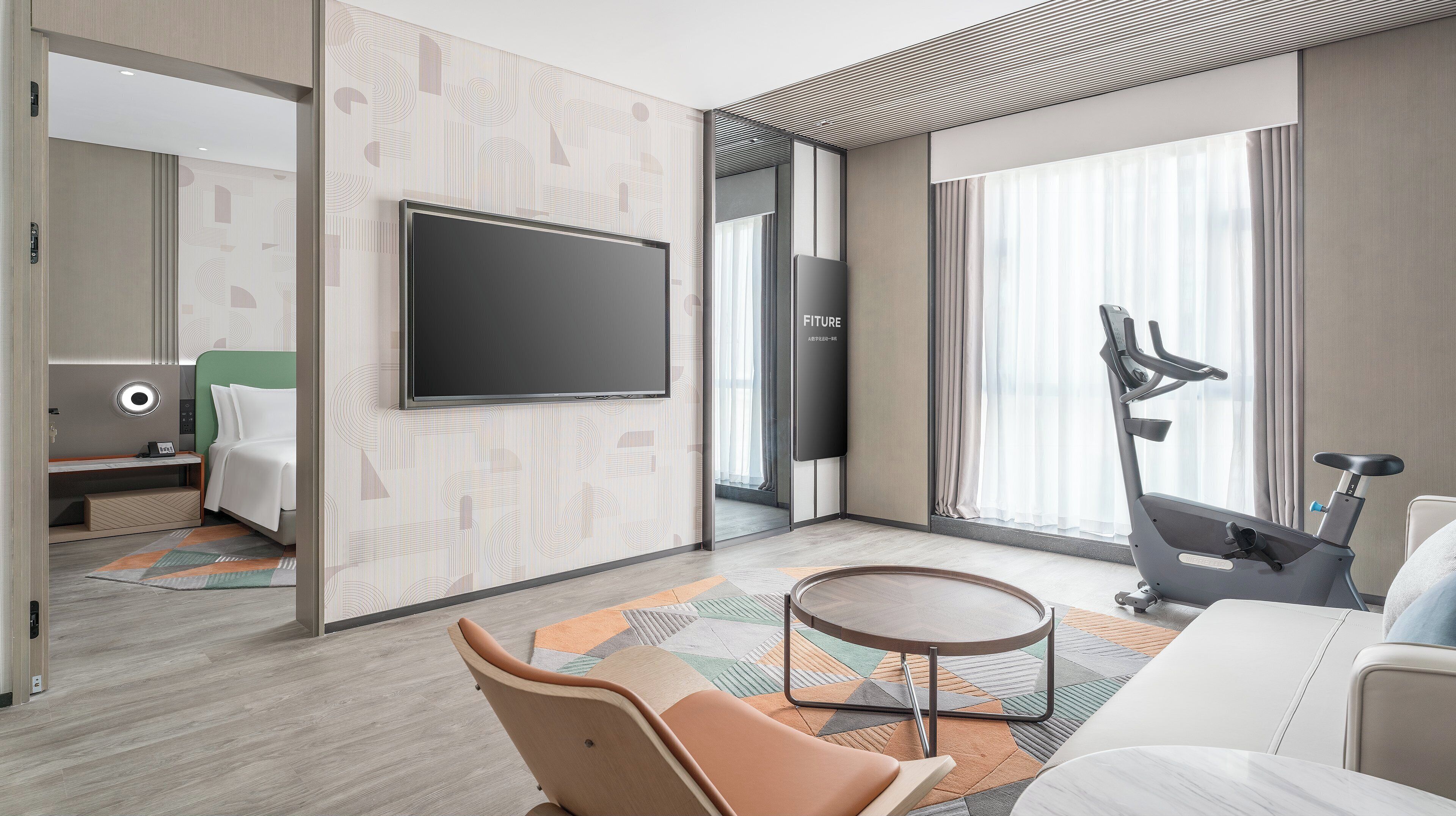A modern hotel room with a large flat-screen TV, exercise bike, and a comfortable seating area with a coffee table.