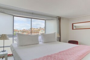 Une chambre d’hôtel avec un grand lit, une fenêtre donnant sur la ville, un fauteuil rouge et un miroir au mur.