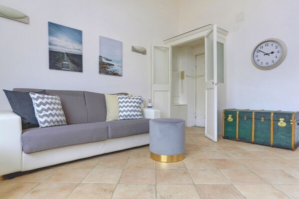 Un salon avec un canapé gris, un pouf gris, une malle verte et une horloge murale.