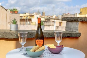 Une bouteille de Prosecco Treviso, deux verres et un bol de collations sont posés sur une table, avec en arrière-plan une vue sur des bâtiments.