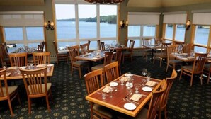 A dining area with multiple tables and chairs, large windows with a view of a body of water, and a patterned carpet.