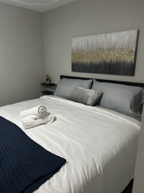 Un lit bien fait, avec une serviette pliée, un plaid bleu foncé et une tête de lit grise.