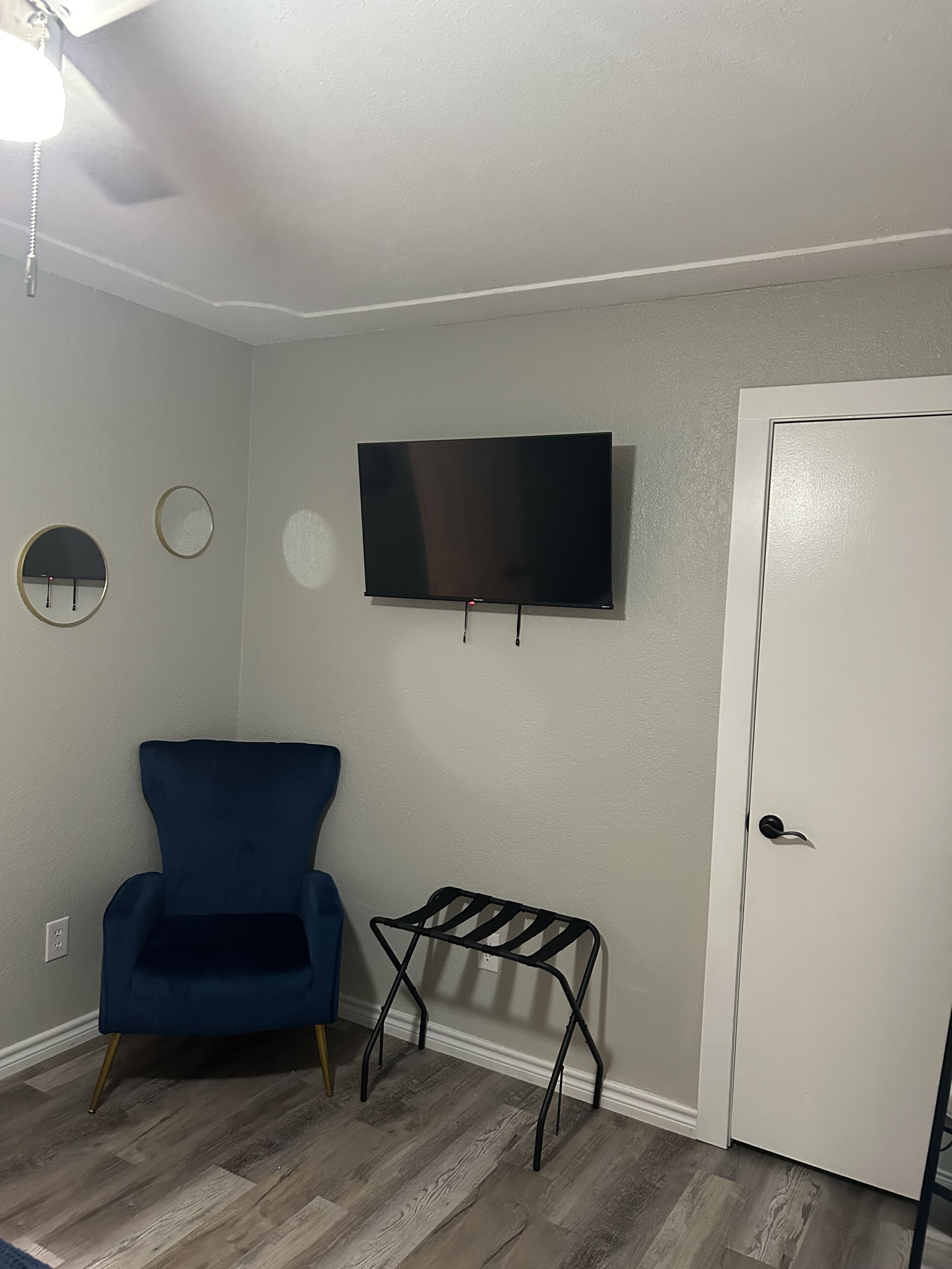 Una habitación con una televisión de pantalla plana montada en la pared, una silla azul, un estante metálico negro pequeño y una puerta blanca.