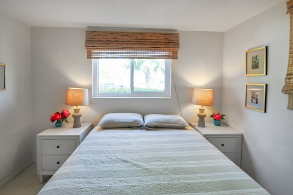 A bedroom with a bed, two bedside tables, two lamps, a window with blinds, and two framed pictures on the wall.