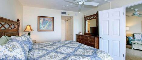Un dormitorio con cama, cómoda, ventilador de techo, un cuadro y una puerta que da a otra habitación.