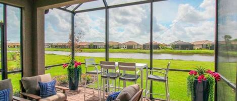 A covered patio with outdoor furniture, a view of a lake, and houses in the background.