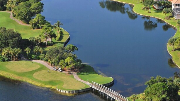 Un campo de golf con un puente sobre una masa de agua, rodeado de árboles y una casa con techo rojo.