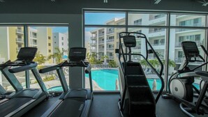 Ein Fitnessraum mit Laufbändern und einem Crosstrainer, großen Fenstern und Blick auf einen Pool sowie Gebäude.