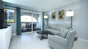 Ein modernes Wohnzimmer mit Sofa, Sessel und Couchtisch. Durch eine Glastür ist ein Essbereich mit Stühlen und Tisch erkennbar.