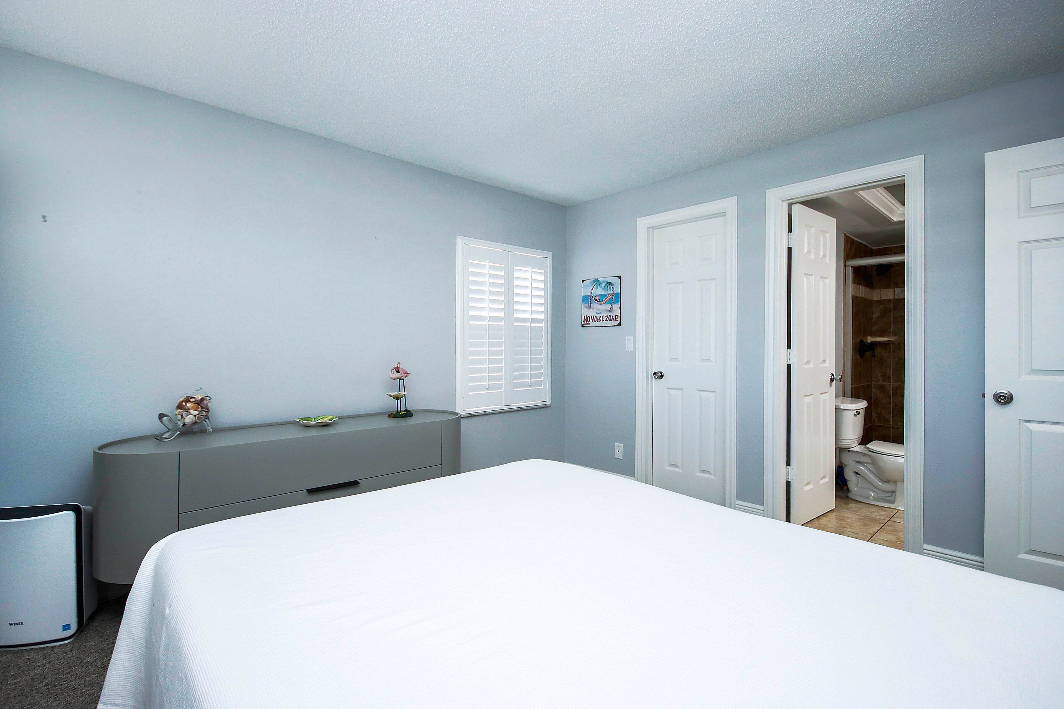 A bedroom with a bed, a dresser, a window with blinds, and a door leading to a bathroom.