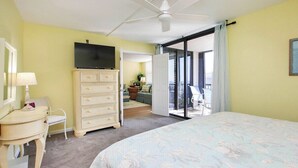 A bedroom with a bed, a dresser, a television, a desk, a chair, and a sliding glass door leading to a balcony.