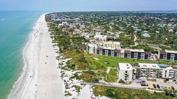 An aerial view of a coastal area with a beach, residential buildings, and a hotel with multiple floors and balconies.