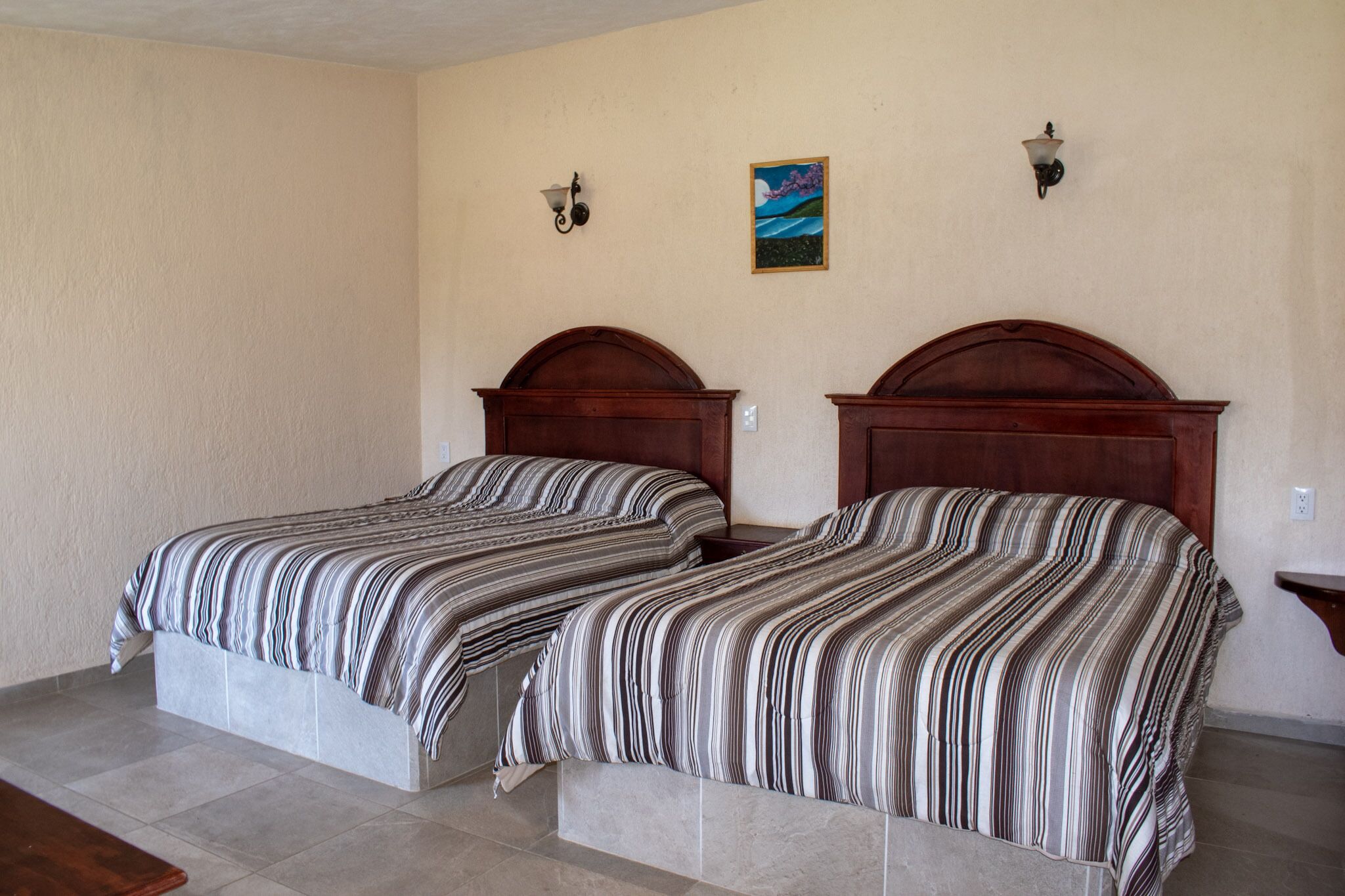Una habitación con dos camas, cada una con cabecero de madera y ropa de cama a rayas. Hay dos lámparas de pared y un cuadro pequeño en la pared.