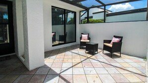 A covered patio area with tiled flooring, two wicker chairs with cushions, and a glass door leading to an interior space.