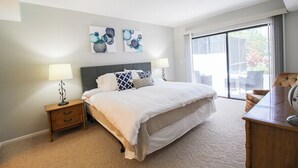 A bedroom with a large bed, two bedside tables with lamps, a desk, and a sliding glass door to a balcony.