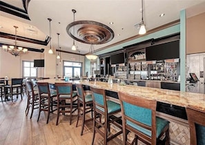 A bar area with a long counter, bar stools, and a television screen. - Second Floor River Strand Condo Overlooking the Golf Course - Sunny Life (Bradenton)