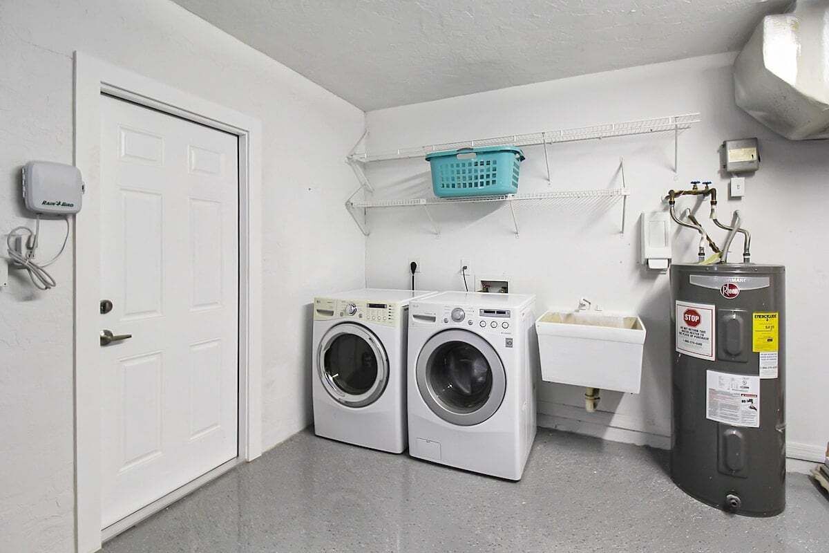 A laundry room with two front-loading washing machines, a sink, and a utility wall with a basket and a water heater.