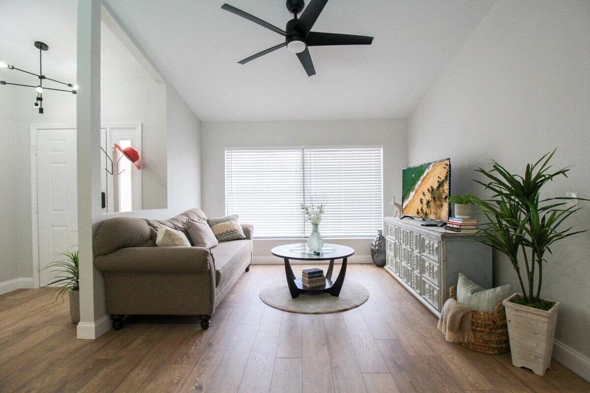 A living room with a sofa, a round coffee table, a ceiling fan, a bookshelf, and a large window with blinds.