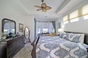 Quarto com cama, penteadeira, espelho, ventoinha no teto, janelas com cortinas e mesa de cabeceira com abajur.