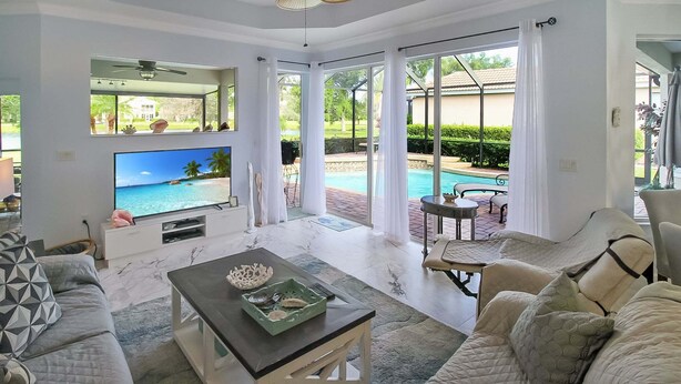 A spacious living room with a large TV, comfortable seating, a ceiling fan, and a view of a swimming pool. - Gorgeous 3/3 private lakefront pool home - Irynas Dream (Bradenton)