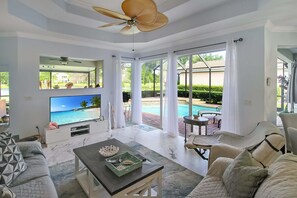 Un salon spacieux doté d’une grande télévision, de sièges confortables, d’un ventilateur de plafond et offrant une vue sur une piscine.