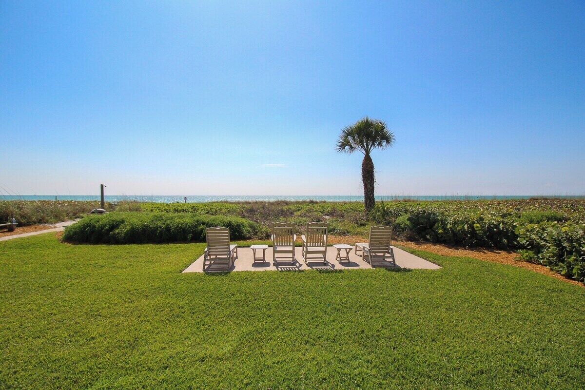 A well-maintained lawn with a seating area featuring chairs and tables, a palm tree, and a view of the ocean.