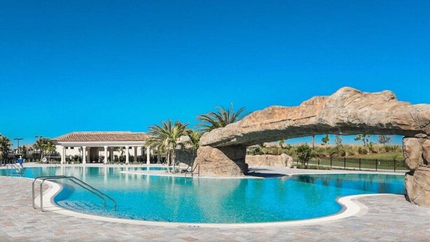 A swimming pool with a natural rock arch, palm trees, and a building in the background. - Exquisite 2 BR / 2 BA Lake Front Condo In Lakewood National Golf Club - Precious Gem (Lakewood Ranch)